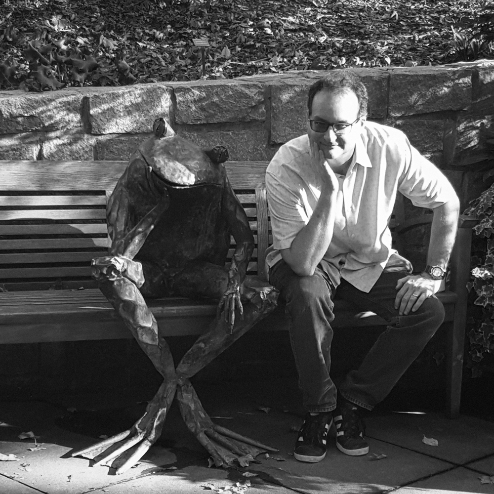 A black and white picture of a man and a frog statue on a bench.