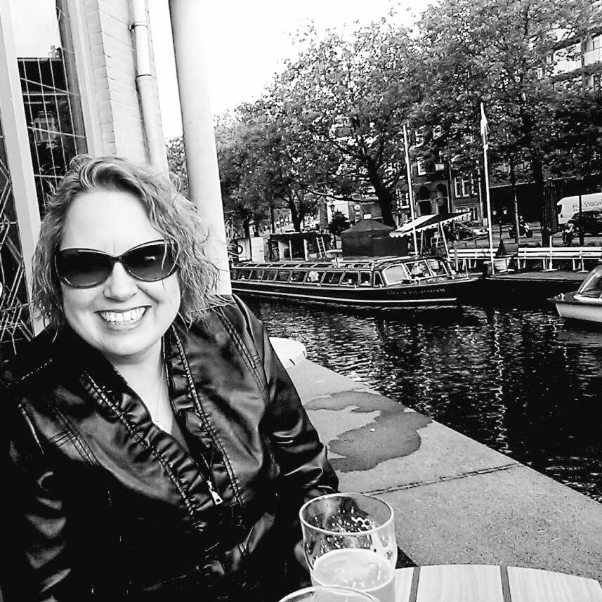 A black and white photo of a woman sitting at a restaurant near some boats in Amsterdam.